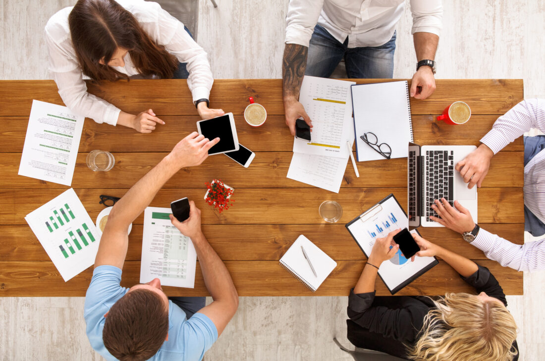 Overhead view of a business team at a wooden table working on laptops, tablets, and smartphones with charts and notes spread out, symbolizing collaboration on B2B tech PR agency pricing strategies.