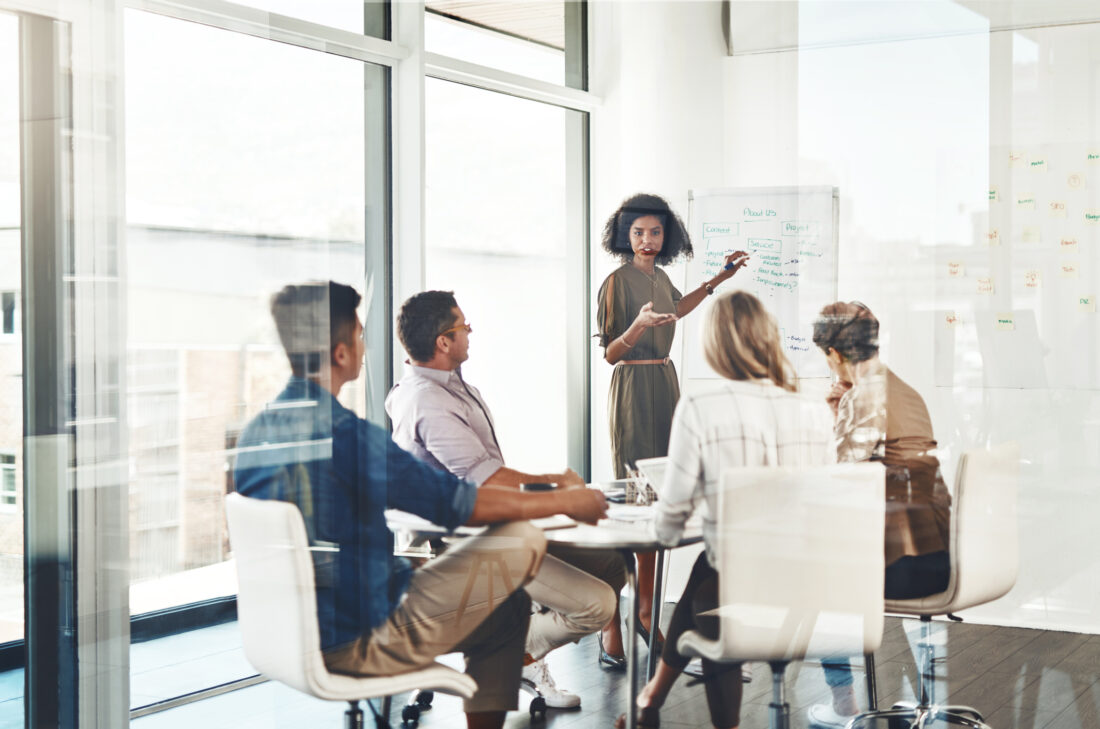 A team of professionals in a modern office listening to a colleague present at a whiteboard, illustrating the benefits of a small PR firm for B2B tech through close collaboration and strategic planning.