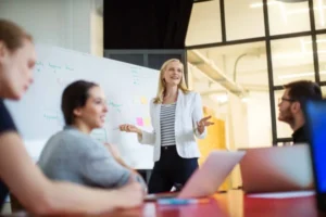 A businesswoman presents to colleagues in a modern office, standing by a whiteboard during a meeting about how to draft an RFP for a PR agency.