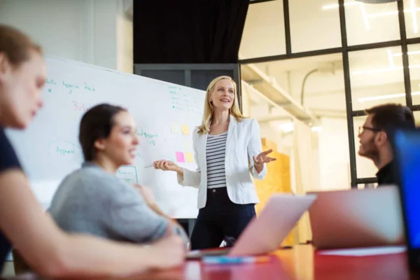 A businesswoman presents to colleagues in a modern office, standing by a whiteboard during a meeting about how to draft an RFP for a PR agency.