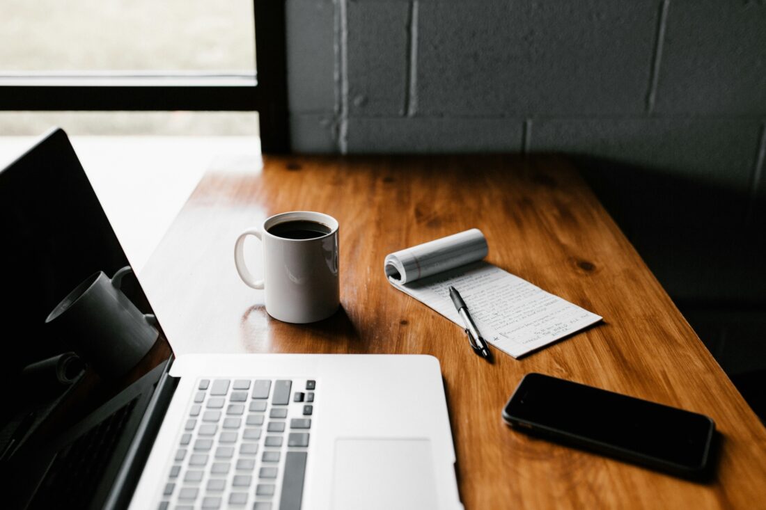 A wooden desk with a laptop, a white coffee mug, a notepad filled with handwritten notes, and a smartphone, symbolizing the process of repurposing good content into new ideas.