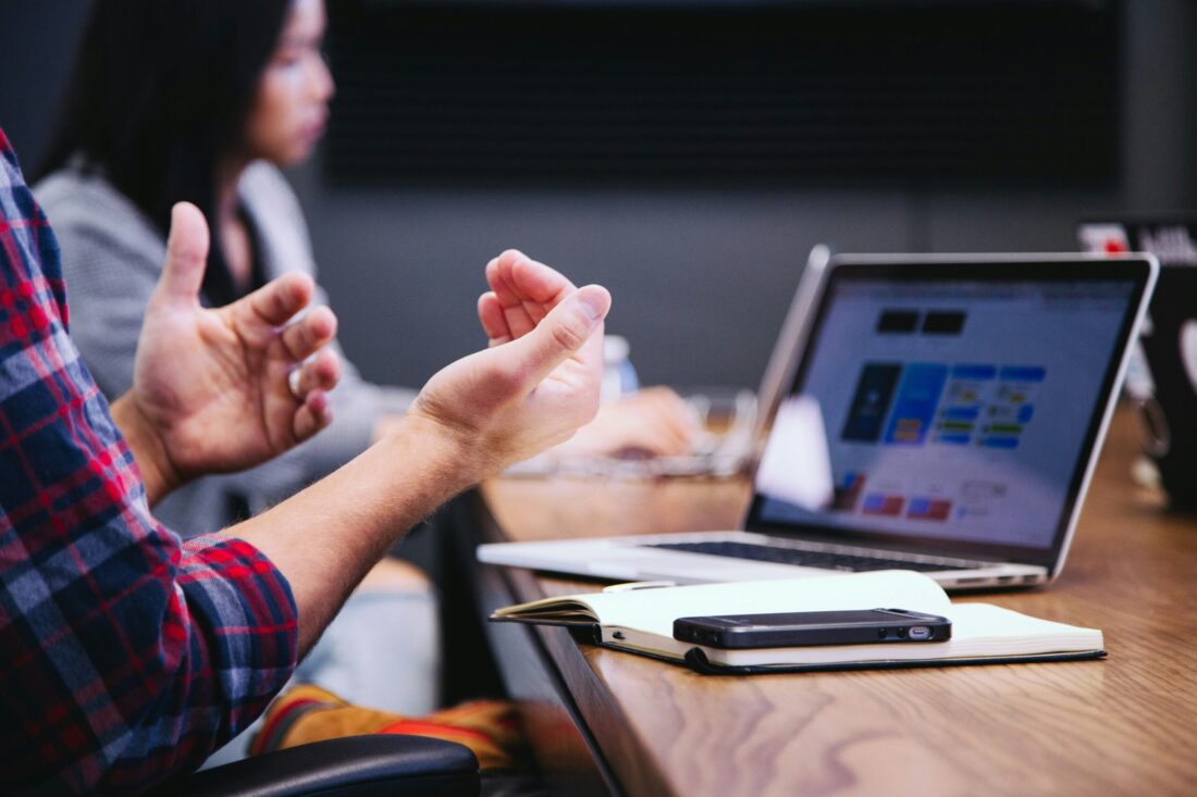 Close-up of a business meeting at a specialized B2B tech PR agency, showing a person gesturing while discussing a digital strategy on a laptop with charts and notes on the table.