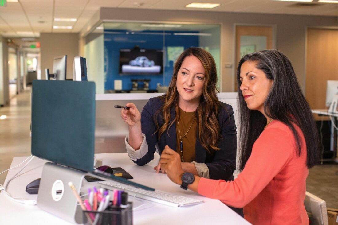 Two colleagues collaborating at a computer in a modern office, discussing whether to hire a PR agency or build an in-house PR team.