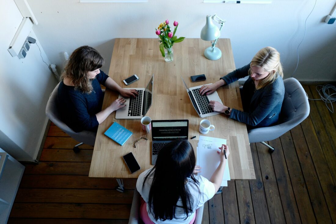 A small PR team collaborates at a wooden table, working on laptops and notes, symbolizing modern “PR for AI visibility” strategies.
